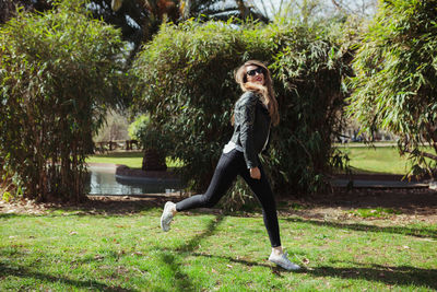 Side view of young woman wearing sunglasses running on grassy field against plants in park