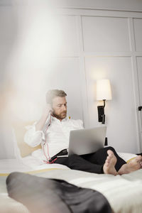 Mature businessman using laptop adjusting headphones while sitting on bed in hotel room