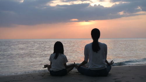 Rear view of men sitting on beach against sky during sunset