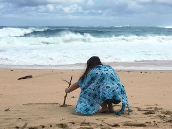 Rear view of woman crouching at beach against cloudy sky