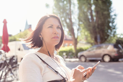 Thoughtful businesswoman looking away while listening music through smart phone on sunny day