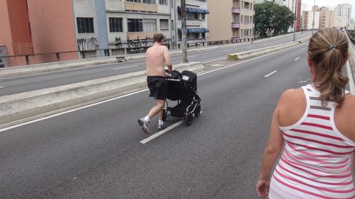 Rear view of shirtless man walking with baby carriage on road in city