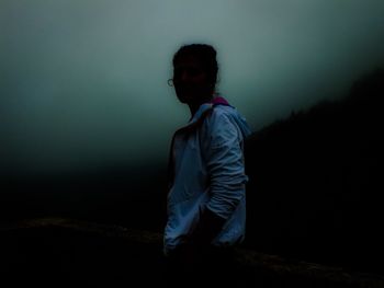 Young man looking away while standing against dark sky