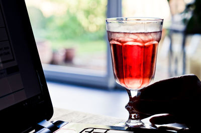 Cropped hand holding glass with red drink by laptop on table 