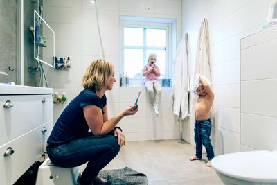 Side view of mother looking at daughter removing top while girl sitting at window in bathroom