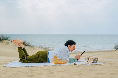 Adult woman on the beach by the ocean in spring sits on a picnic, reads a book on a tablet, watches