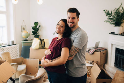 Smiling man embracing pregnant woman while standing against boxes in living room