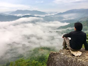 Full length of hiker sitting on rock by mountains against cloudy sky