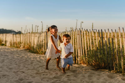 Side view of cheerful siblings standing on sand near wooden fence while observing residential houses near mountainous area