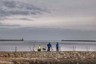 People fishing on retaining wall against sky during sunset