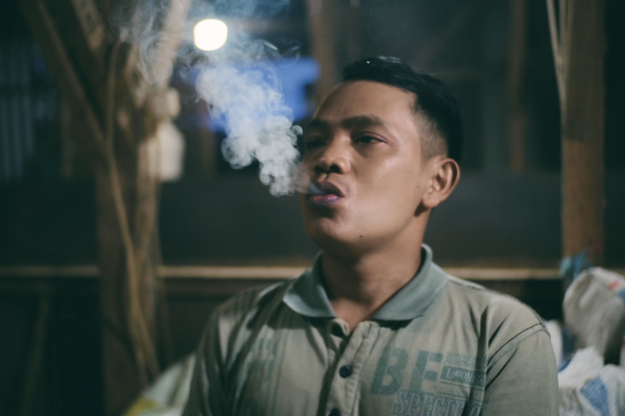 one person, smoke, portrait, headshot, smoking, adult, men, young adult, social issues, smoking issues, indoors, looking away, night, focus on foreground, looking, person, front view, casual clothing, contemplation, blue, cigarette, bad habit, activity