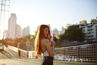 Side view of beautiful young woman standing on street in city against sky during sunset
