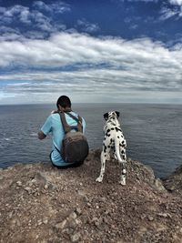 Rear view of man with backpack by dog sitting against sea and cloudy sky