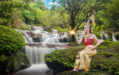 Beautiful young woman wearing crown and traditional clothing while sitting against waterfall in forest