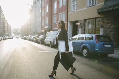 Side view of female realtor carrying signboard and bag while crossing street in city