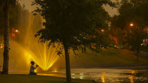 Woman standing by illuminated tree by lake against sky during sunset