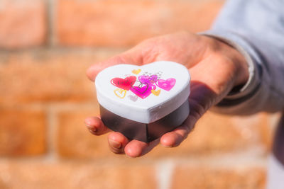 Cropped hand of man holding heart shape gift box against brick wall