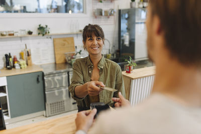 Smiling female owner giving back credit card to male customer at checkout in coffee shop