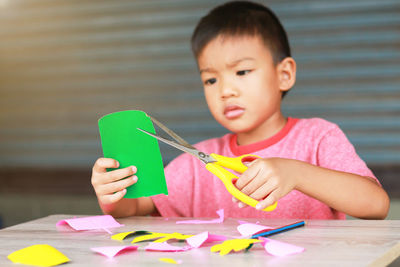 Boy cutting green paper while sitting against corrugated iron