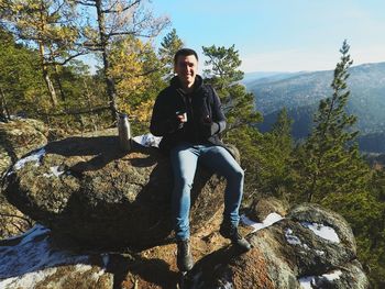 Portrait of young man sitting on rock against sky