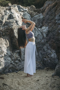 Side view of ethnic female with praying hands practicing yoga on sandy land against rock in sunlight