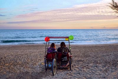 Rear view of women sitting on pedicab at beach against sky during sunset