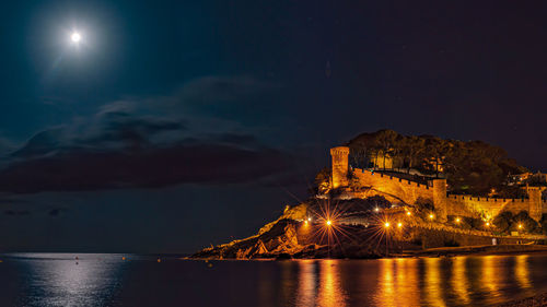 Illuminated buildings by sea against sky at night