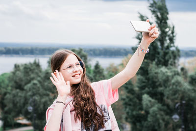 A cheerful girl in glasses greets by video call on a smartphone on the background of a river 