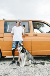 Young man with dog wearing leg prosthesis and using cell phone at camper van
