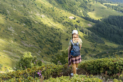 Rear view of young woman with backpack hiking in mountains among rhododendron plants and flowers 