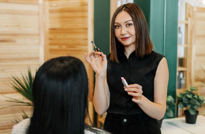 A make-up artist makes makeup for a girl in the salon, holds a lipstick and a brush in her hand.