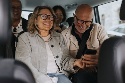 Happy male and female senior friends sharing smart phone while sitting in car