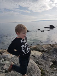 Boy looking away while standing with hand on hip at rocky shore