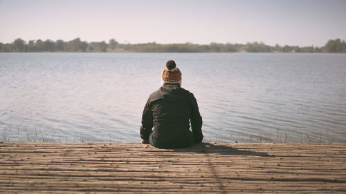 Rear view of man sitting on pier over lake against sky