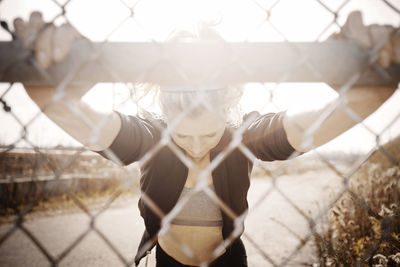 Athlete stretching in front of fence against road on sunny day