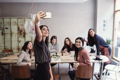 Smiling young woman taking selfie with female coworkers at table in workshop