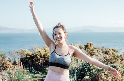 Side view of young woman with arms raised standing at beach against sky