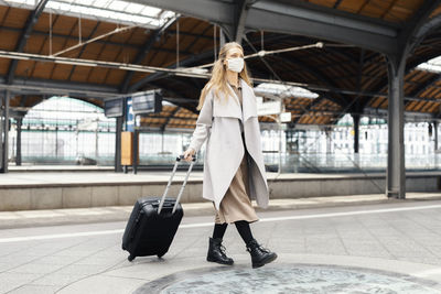 Woman wearing mask walking with suitcase at railroad station