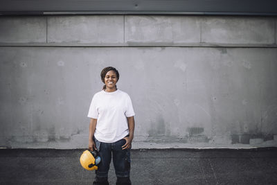 Portrait of smiling female construction worker with hand in pocket standing on road against wall