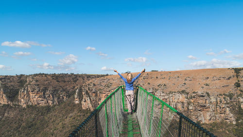 Rear view of woman with arms outstretched on footbridge against blue sky