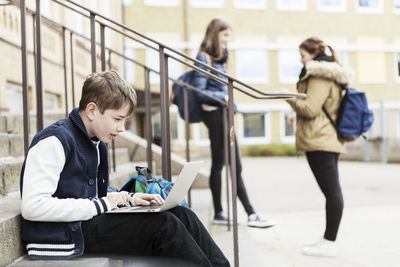 Boy using laptop on steps with schoolgirls in background outside school building