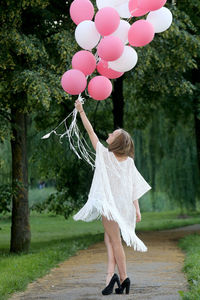 Woman holding pink and white helium balloons on footpath against trees at park