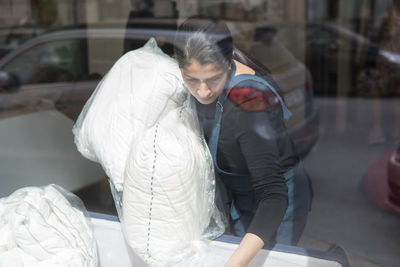 Mature female dry cleaner carrying pillows seen from laundromat window