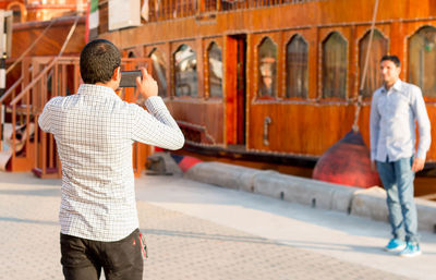 Man photographing friend standing against wooden building