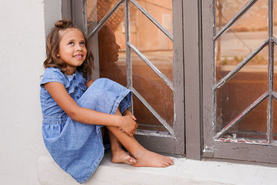 Cute girl looking away while sitting on window sill