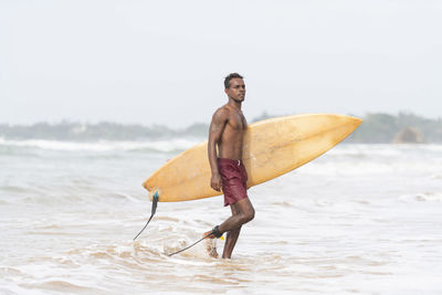 Shirtless man with surfboard wading in sea against sky