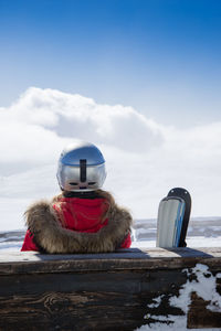 Rear view of person with snowboard sitting on bench against sky