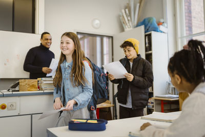 Smiling male and female students walking in classroom holding documents