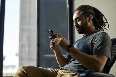 Side view of businessman with dreadlocks using smart phone while sitting on chair against window in office