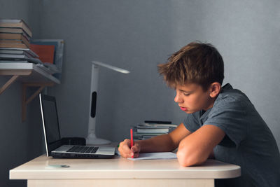 Student teenager boy doing homework with laptop, open copybook and computer, workplace at home